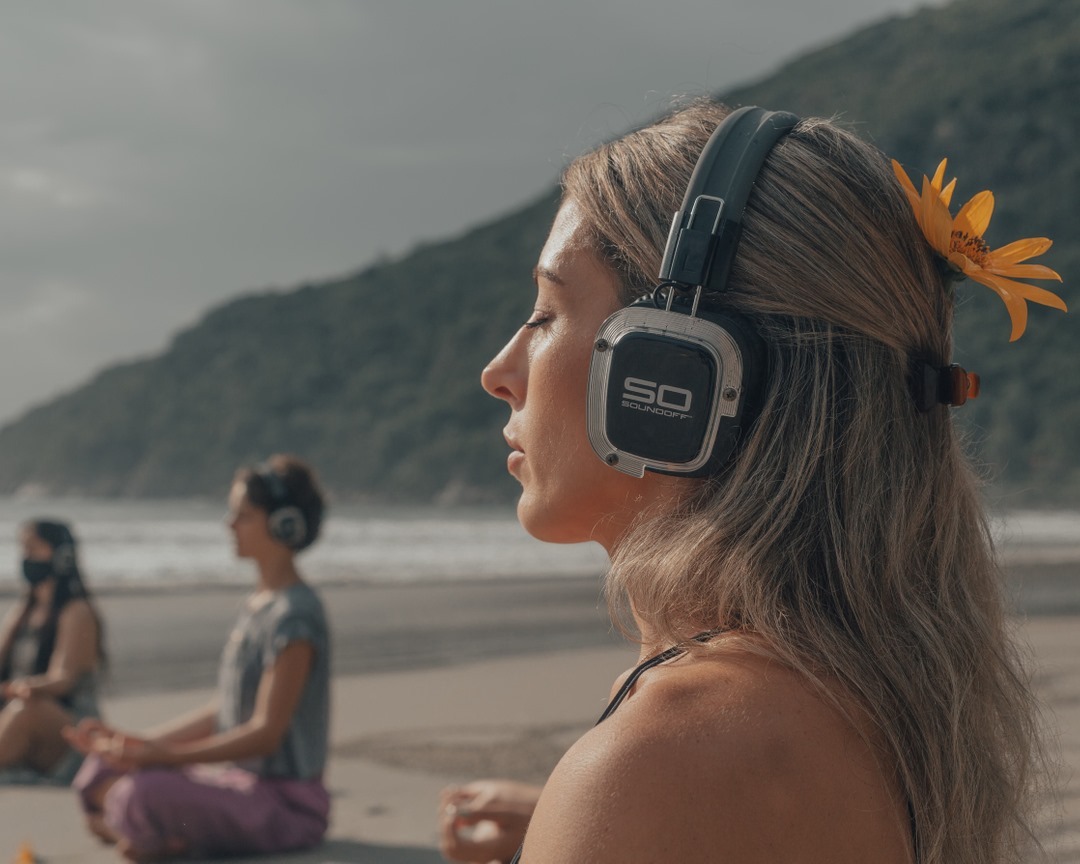 Woman with flower in her hair meditating while wearing Sound Off headphones on a beach